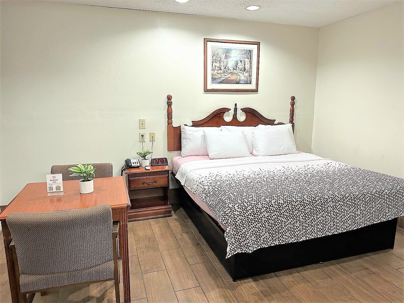 Habitación de hotel con cama, mesita de noche de madera, una silla, una mesa y un cuadro en la pared.