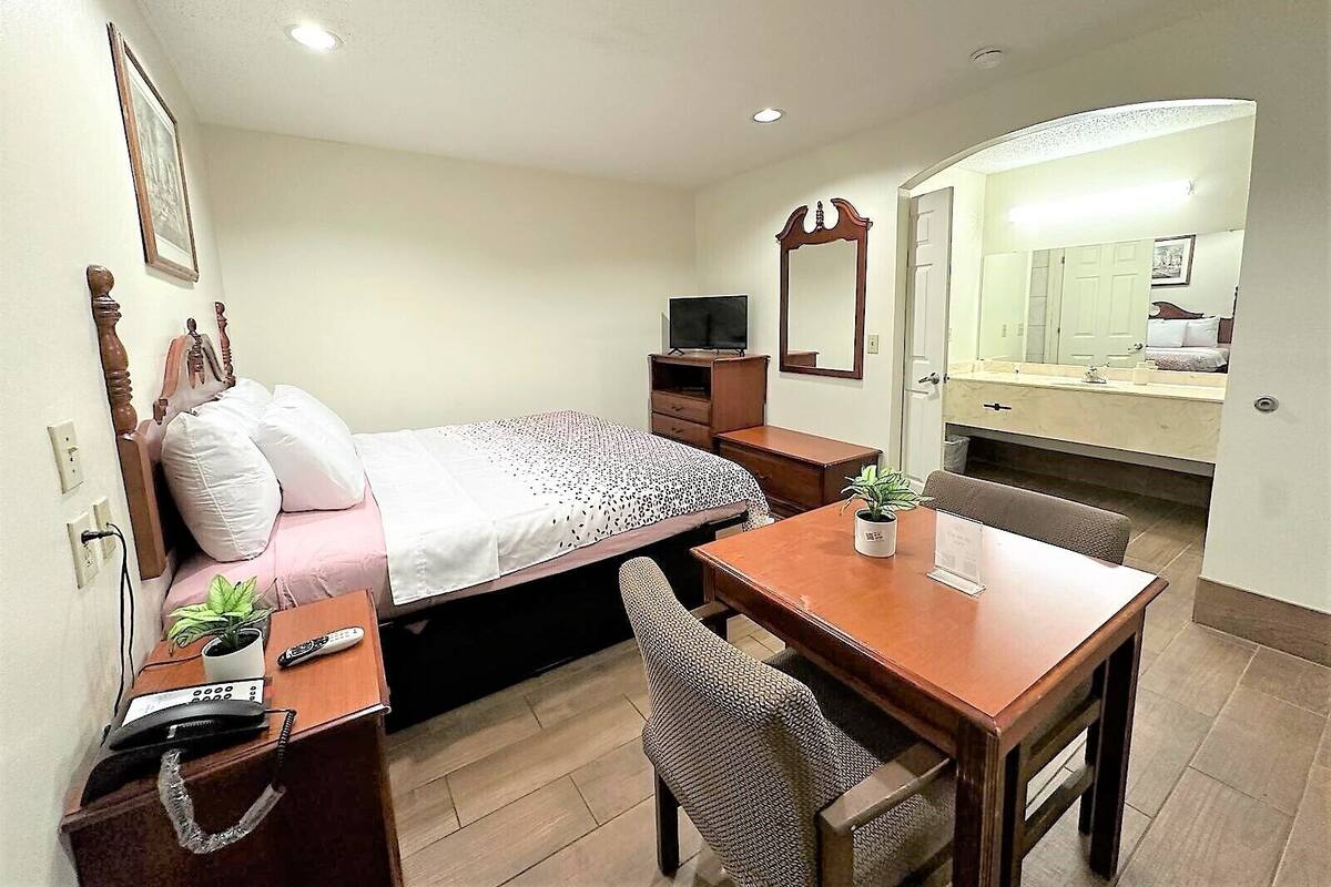 A hotel room with a bed, a desk with a chair, a television, a mirror, and a bathroom visible through an open door.