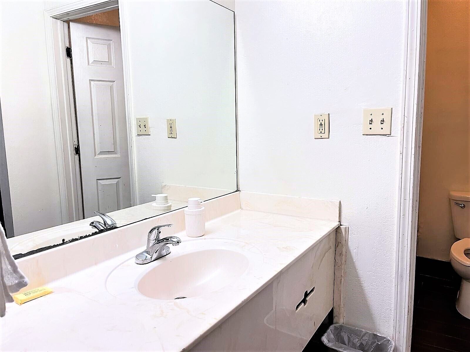 Un baño con un lavamanos blanco, un espejo y un inodoro. Hay dos enchufes en la pared.
