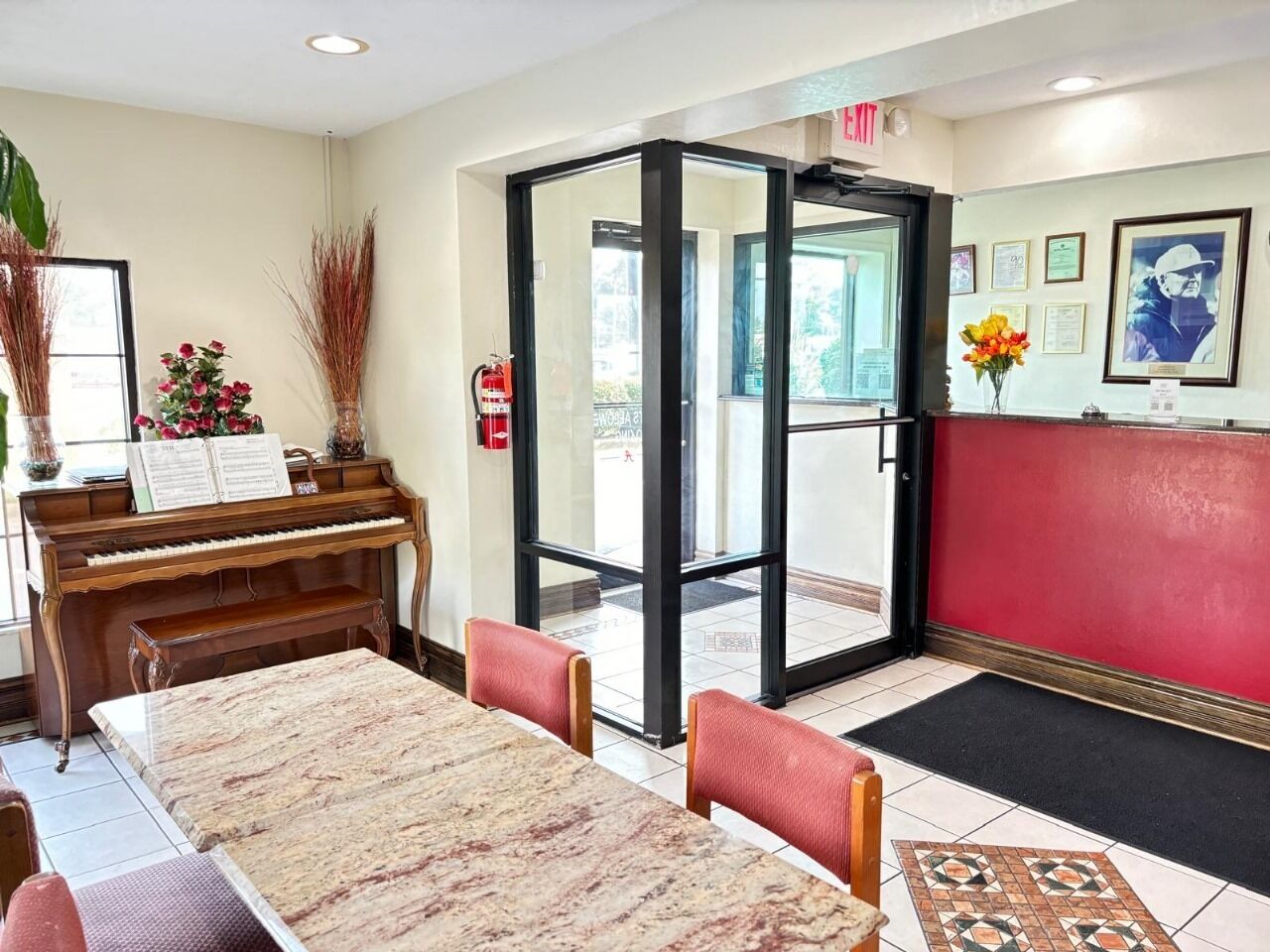Un vestíbulo con piano, una mesa de mármol, sillas rojas, una entrada por puerta de cristal y una pared roja con cuadros enmarcados.