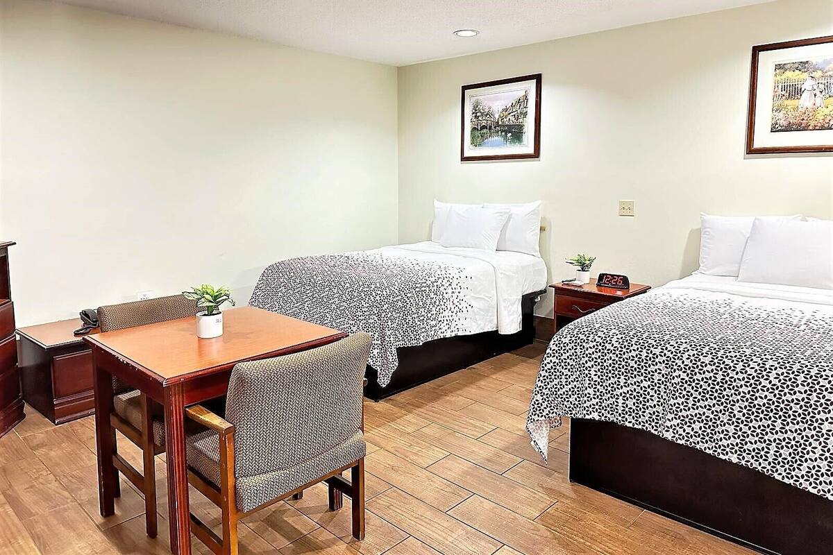A hotel room with two beds, a wooden desk, a chair, a small table with a plant, and a clock on the bedside table.