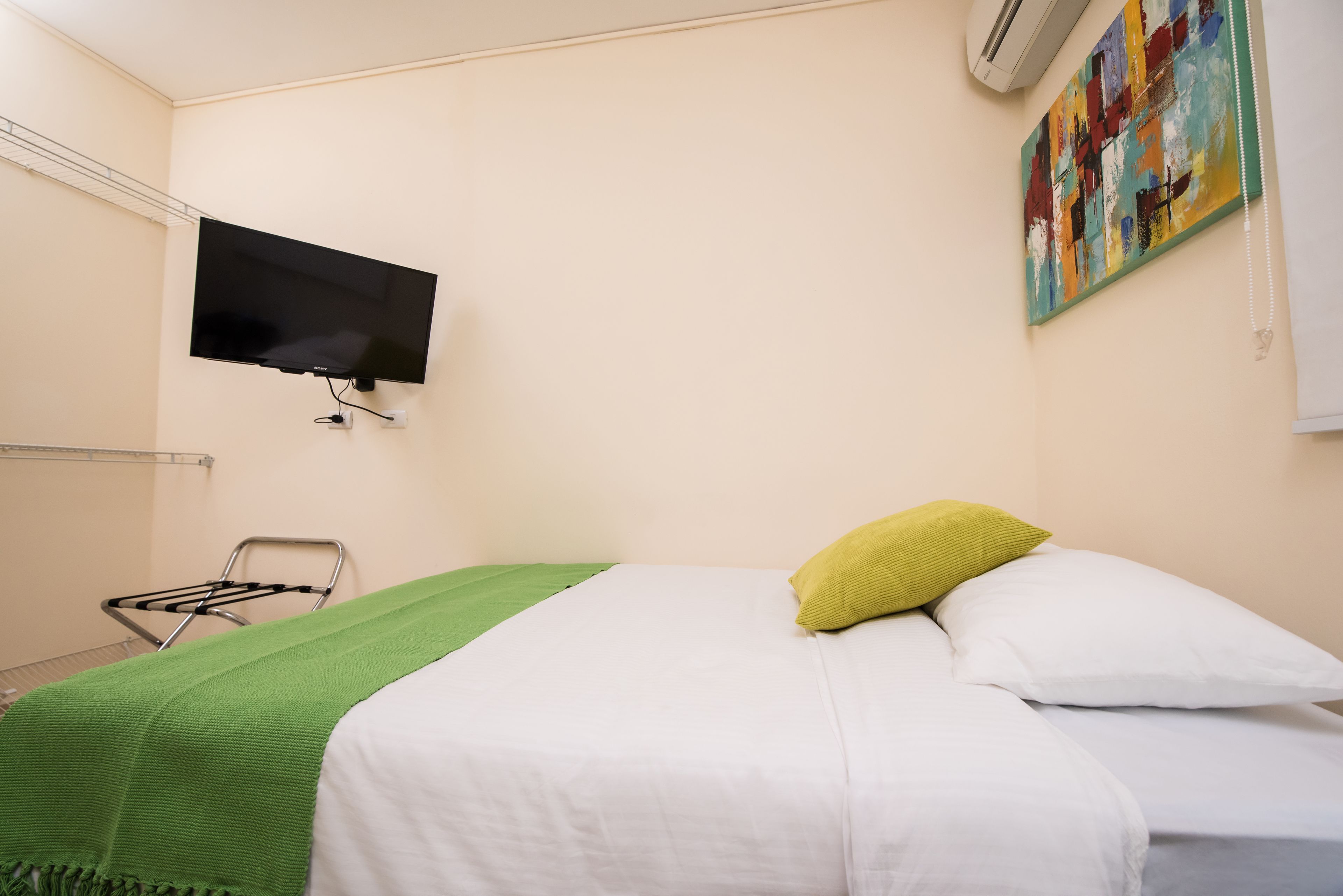 A hotel room with a bed, a green blanket, a white pillow, a TV mounted on the wall, and a colorful painting.