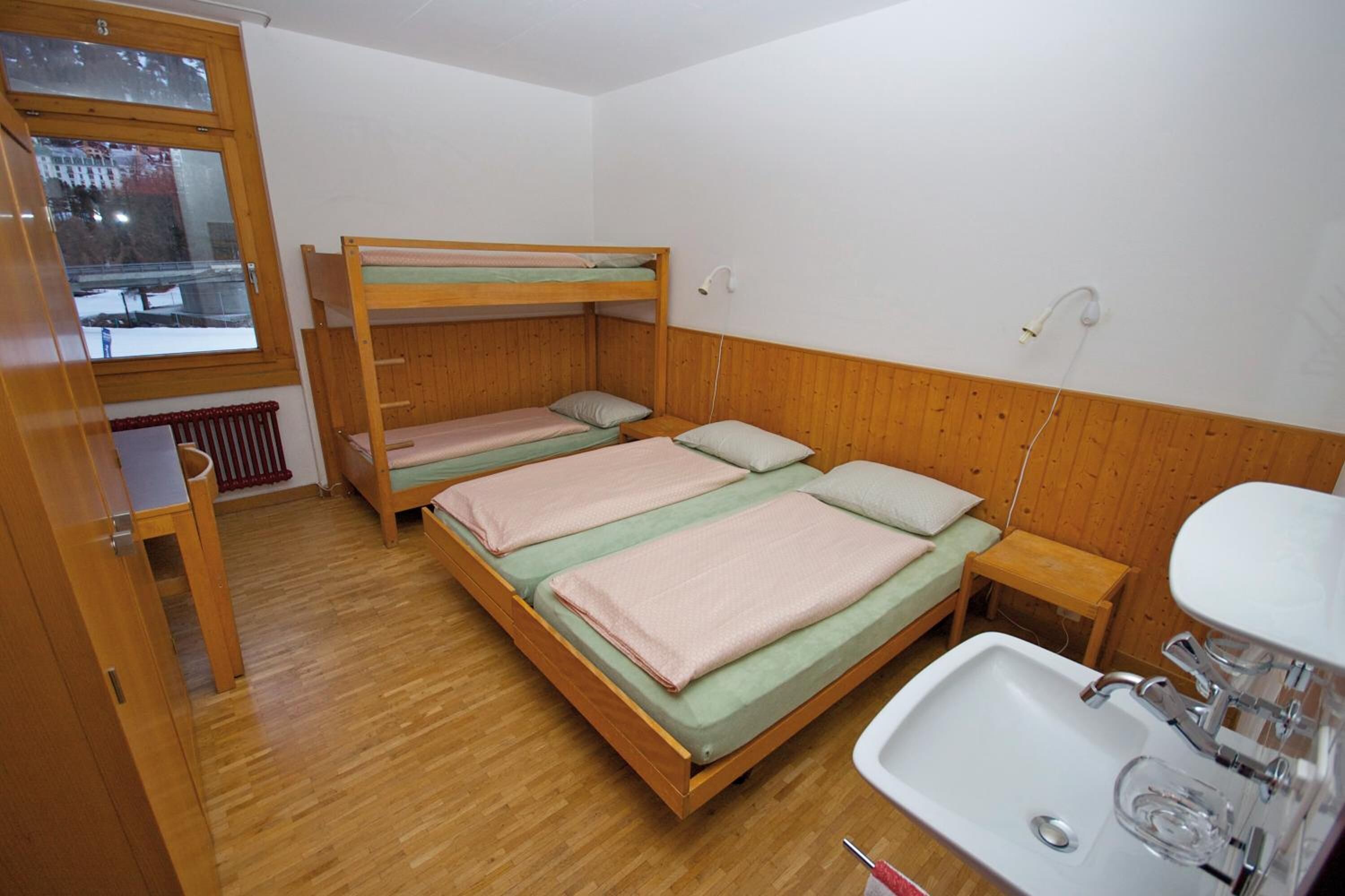 A small, clean room with a bunk bed, a wooden floor, a window, and a sink.