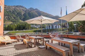 Un espace extérieur aménagé avec des tables et des chaises, des parasols, et une vue sur les montagnes et les bâtiments en arrière-plan.