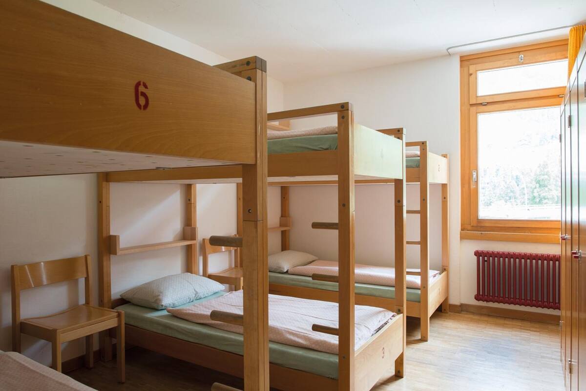 Une chambre avec trois lits superposés, une chaise en bois, une petite table et une fenêtre donnant sur les arbres.