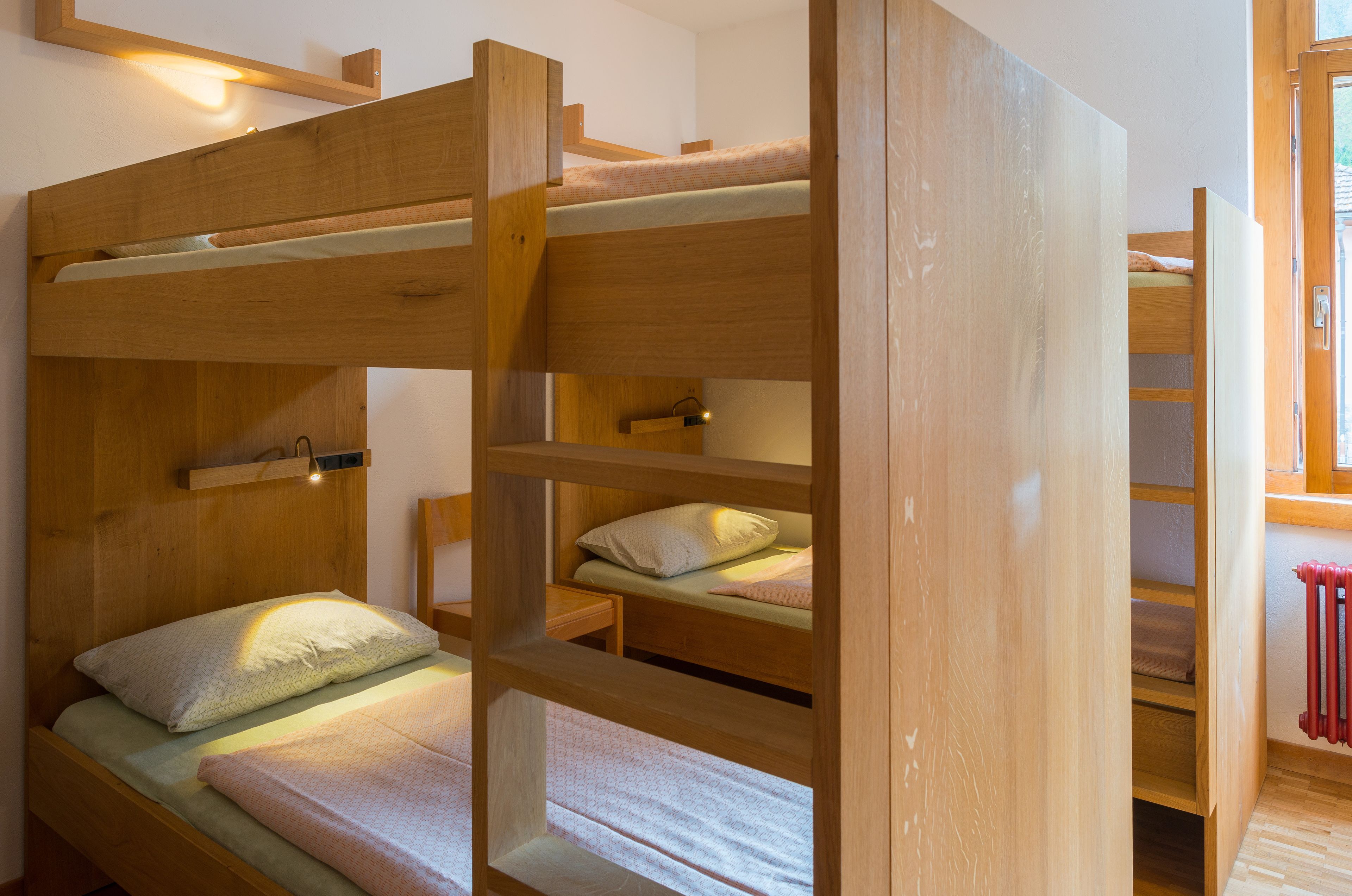 A bunk bed room with wooden construction, a small bedside table, and a light fixture above the top bunk.