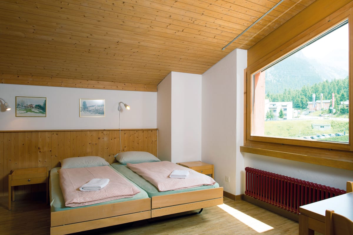 Une chambre avec un plafond en bois, une grande fenêtre donnant sur les montagnes, un lit avec du linge de lit rose et une table de chevet.