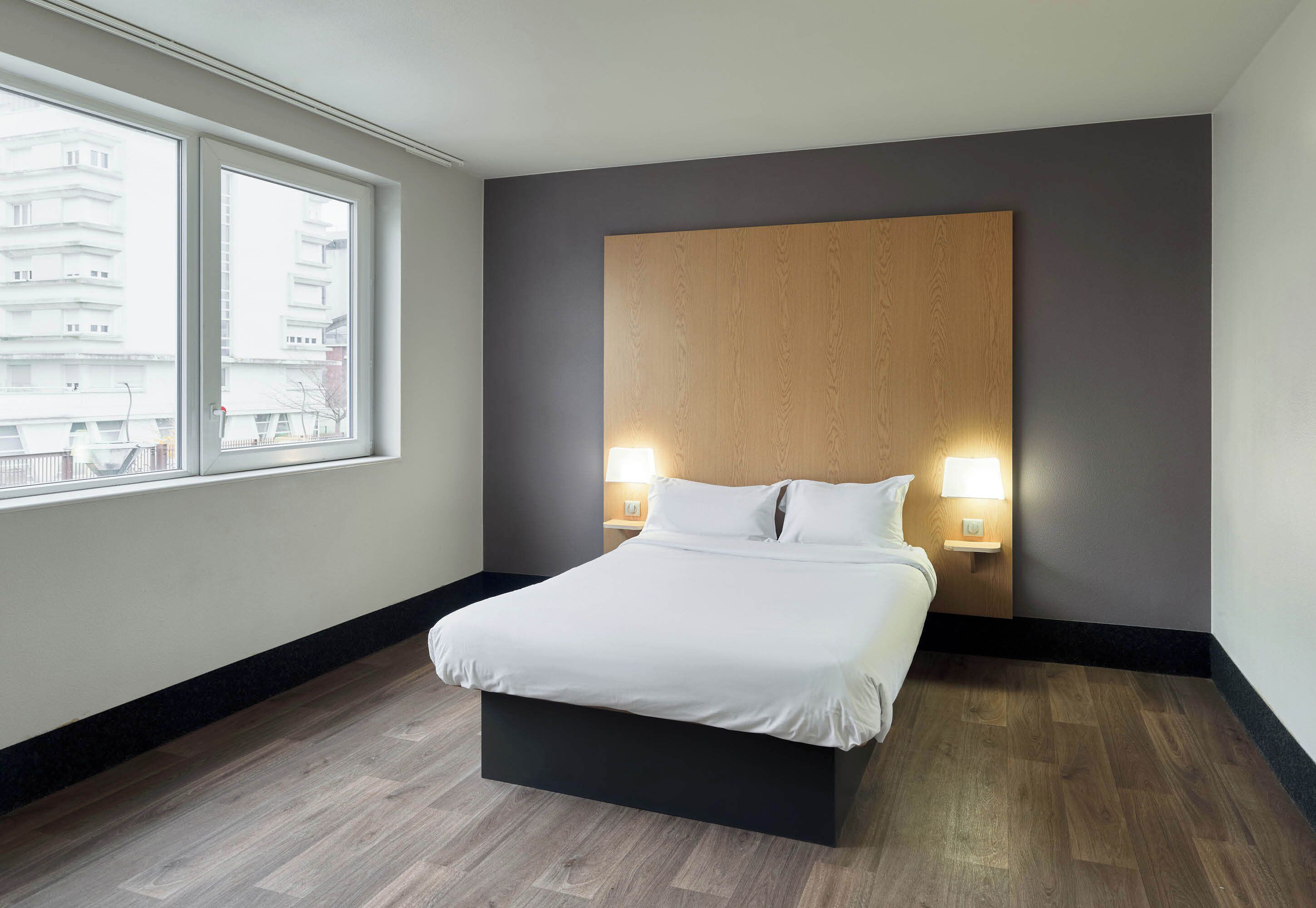 Ein modernes Hotelzimmer mit einem großen Bett, zwei Nachttischlampen, einem Holz-Kopfteil und Blick auf Gebäude durch ein großes Fenster.