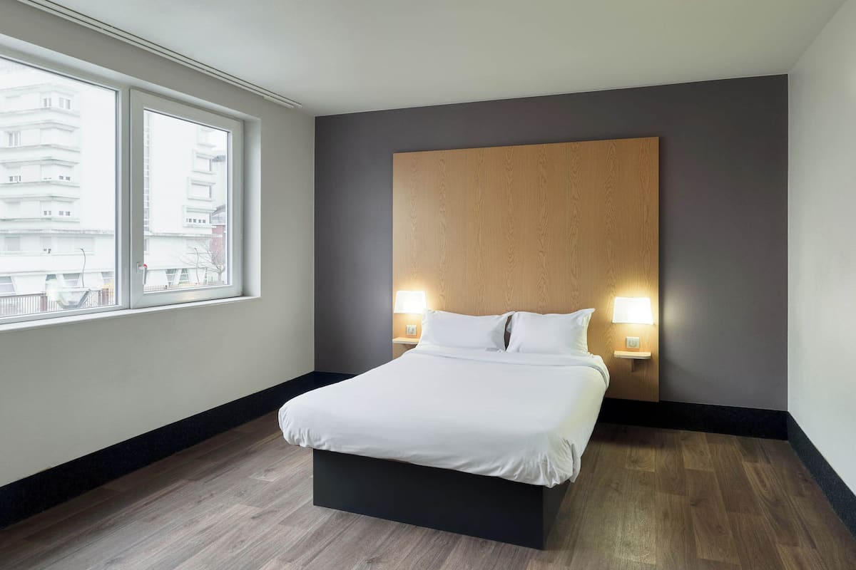 A modern hotel room with a large bed, two bedside lamps, a wooden headboard, and a view of buildings through a large window.