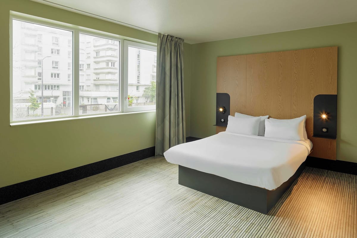 A hotel room with a large bed, a view of buildings through the window, and a wooden headboard.