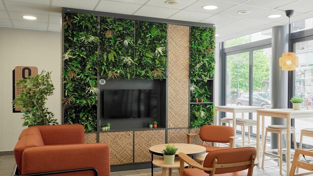A modern interior with orange seating, a round table, a TV screen, and a green wall with plants.