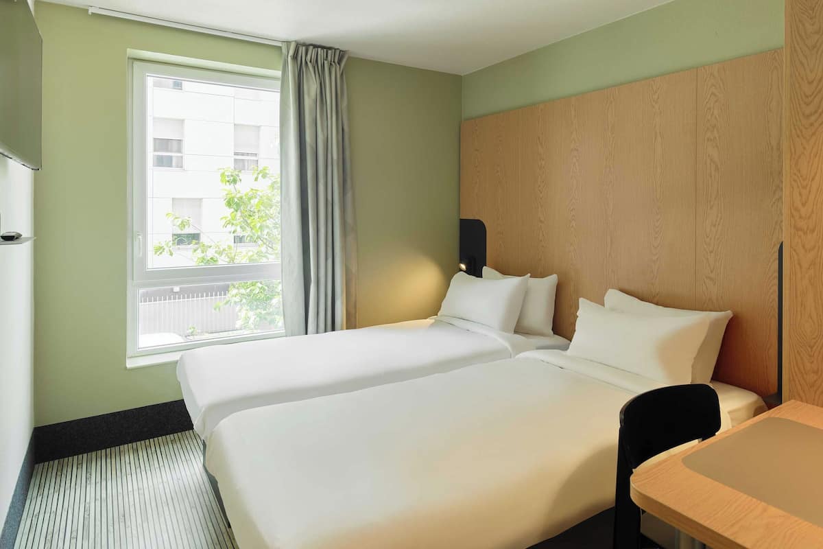 A hotel room with two beds, a wooden headboard, a window with a view of buildings, and a small table.