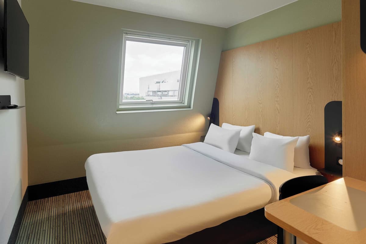 A hotel room with a large bed, a small desk, a window with a view of a building, and a TV mounted on the wall.