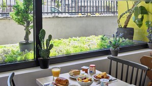 A breakfast table with croissants, fruit, coffee, and juice. There are potted plants and a view of greenery outside.