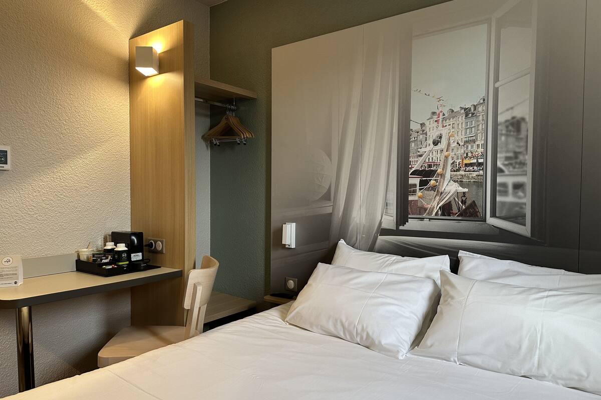 A hotel room with a bed, a desk with a coffee maker, a chair, and a window with a cityscape view.