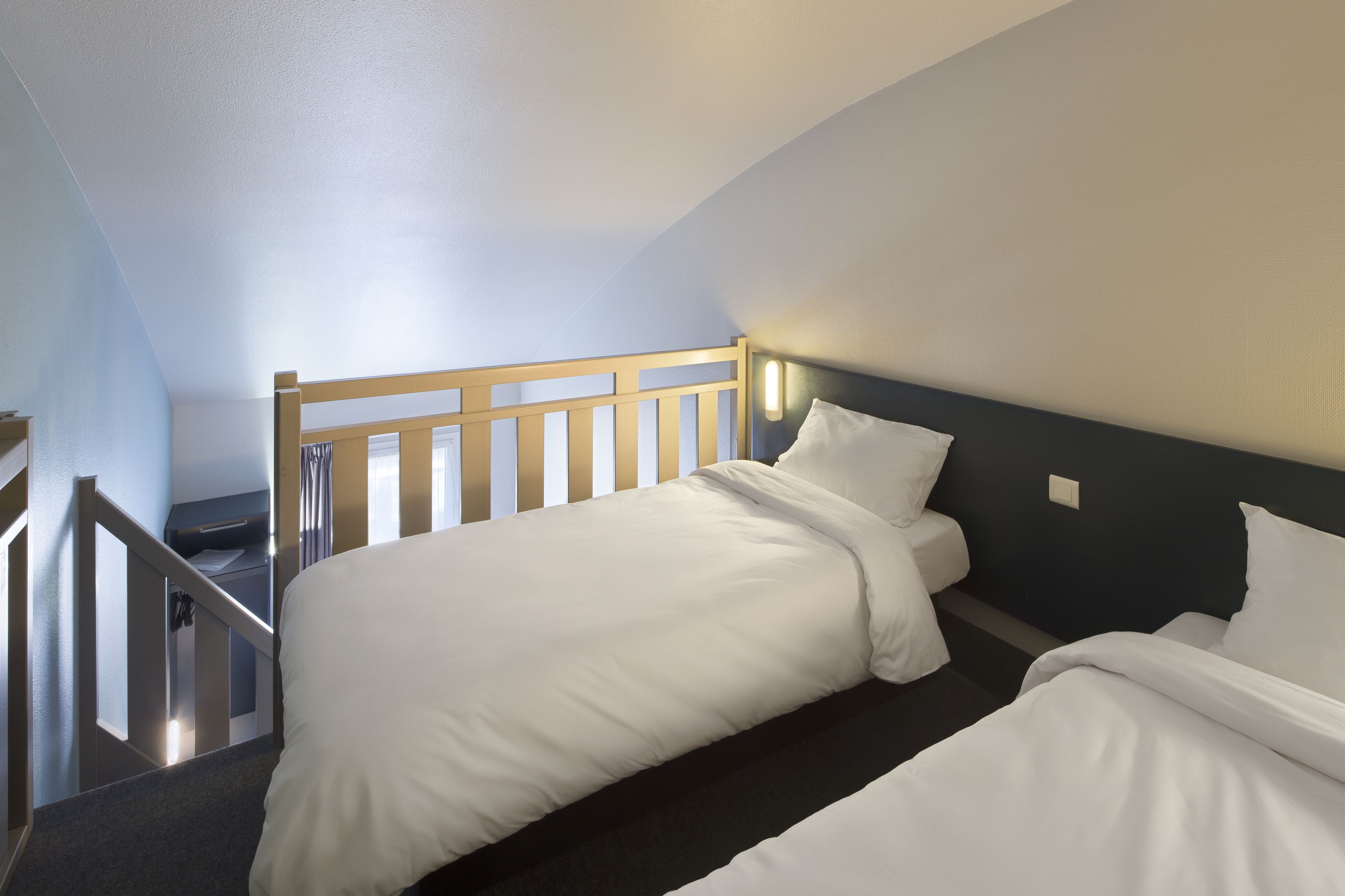 A hotel room with two single beds, a wooden headboard, a dark wall, and a staircase leading to an upper level.