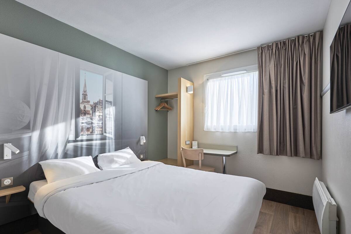 A hotel room with a bed, a desk, a chair, a window with curtains, and a view of a cityscape.