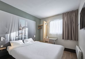 A hotel room with a bed, a desk, a chair, a coat rack, and a window with curtains.