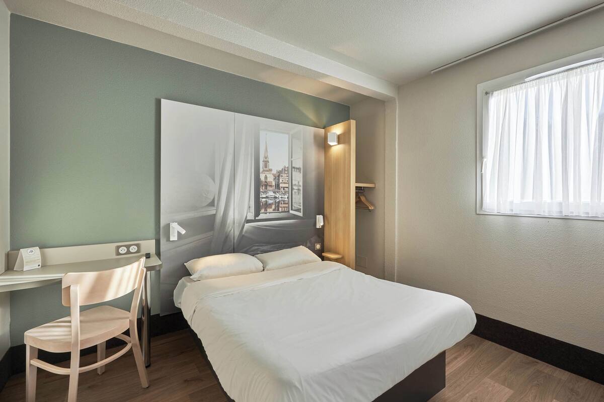 A hotel room with a bed, a desk, and a chair. There is a window with curtains, a wall-mounted light, and a view of a cityscape through the window.