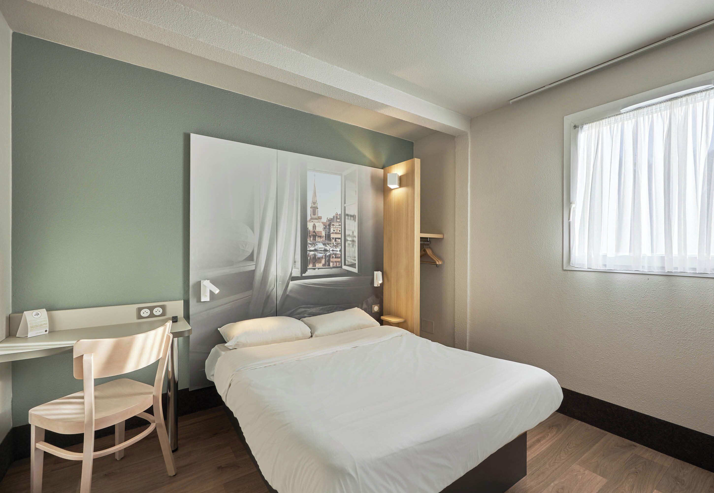 Quarto de hotel com uma cama, uma escrivaninha, uma cadeira, uma janela e vista para a paisagem urbana.