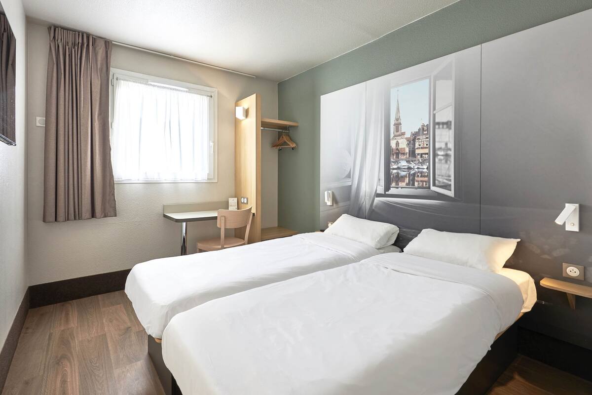 A hotel room with two beds, a desk, a chair, and a large window with a cityscape view.