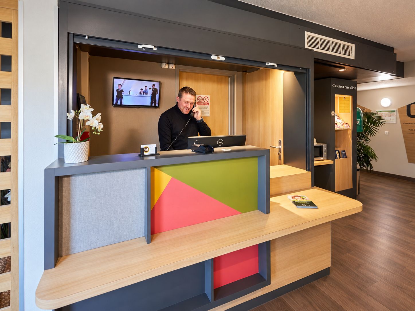 A reception desk with a computer, a phone, and a monitor displaying a video. There is a plant on the desk, and a sign with a prohibition symbol.