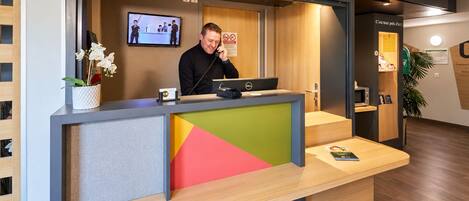 A reception desk with a computer, a phone, and a monitor displaying a video. There is a plant on the desk, and a sign with a prohibition symbol.