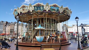 Un carrusel con decoraciones ornamentales y caballos, rodeado de bancos y faroles, situado en una plaza pública con edificios al fondo.