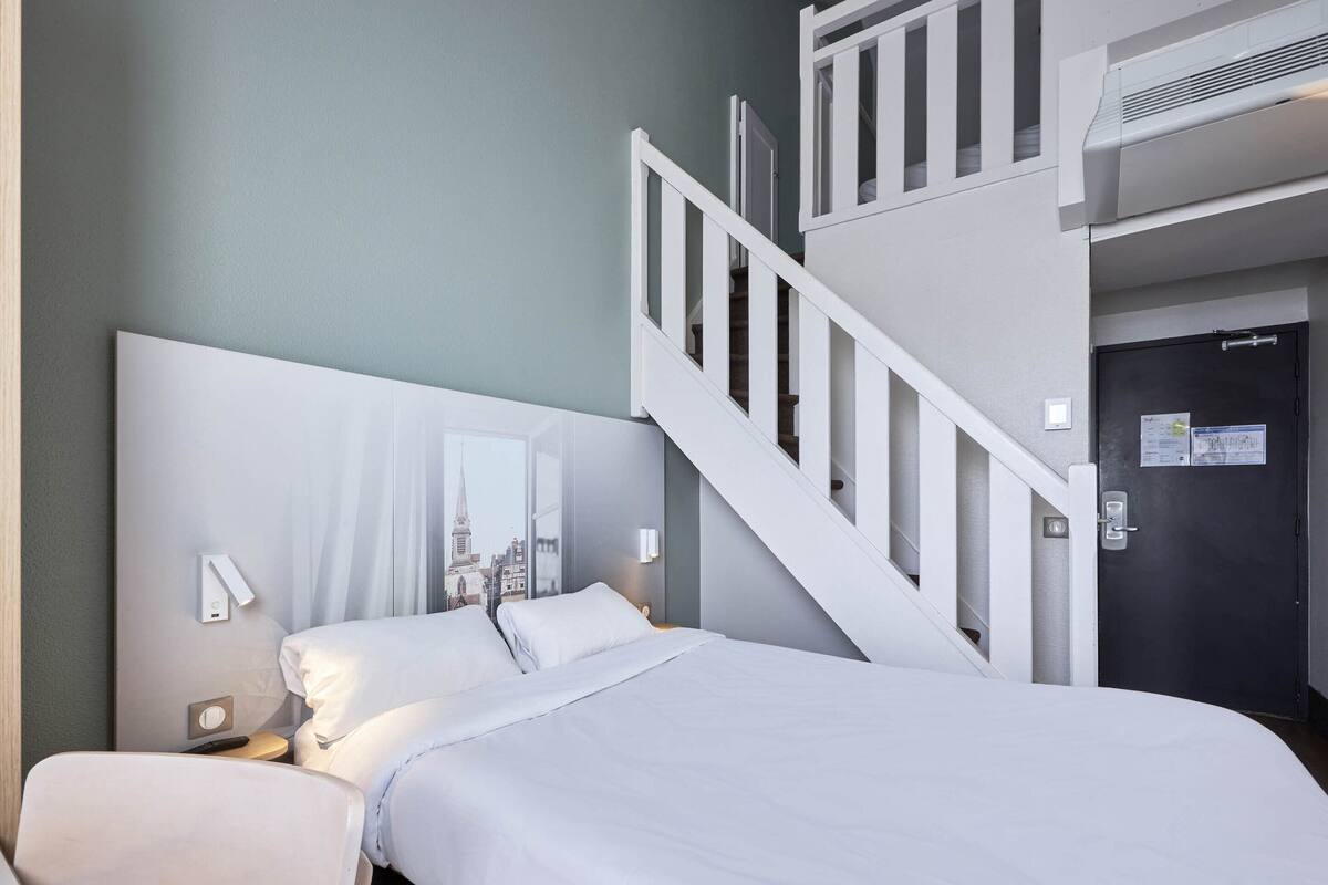 A hotel room with a bed, a nightstand, a wall-mounted mirror, a staircase, and a door with a lock.
