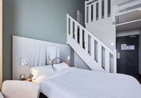 Habitación de hotel con cama, mesita de noche, espejo en la pared, una escalera y una puerta con cerradura.