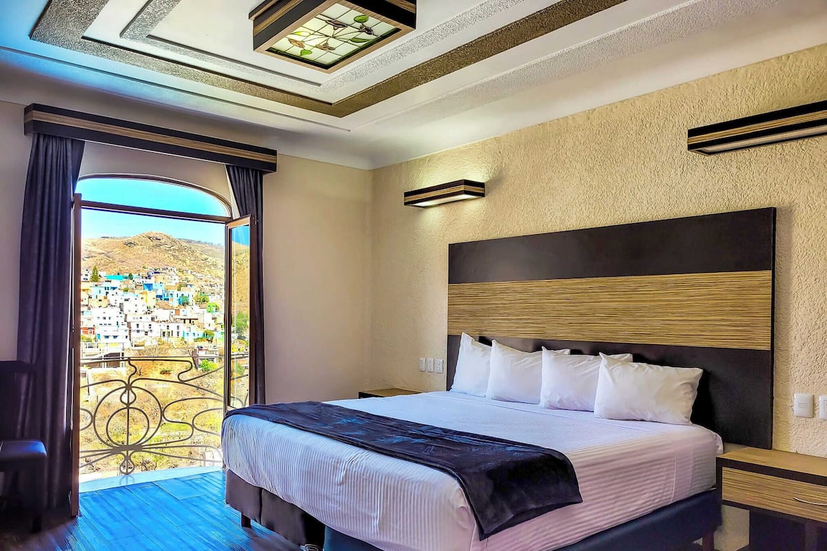 A hotel room with a large bed, a nightstand, a view of a town through a balcony door, and a ceiling with a skylight.