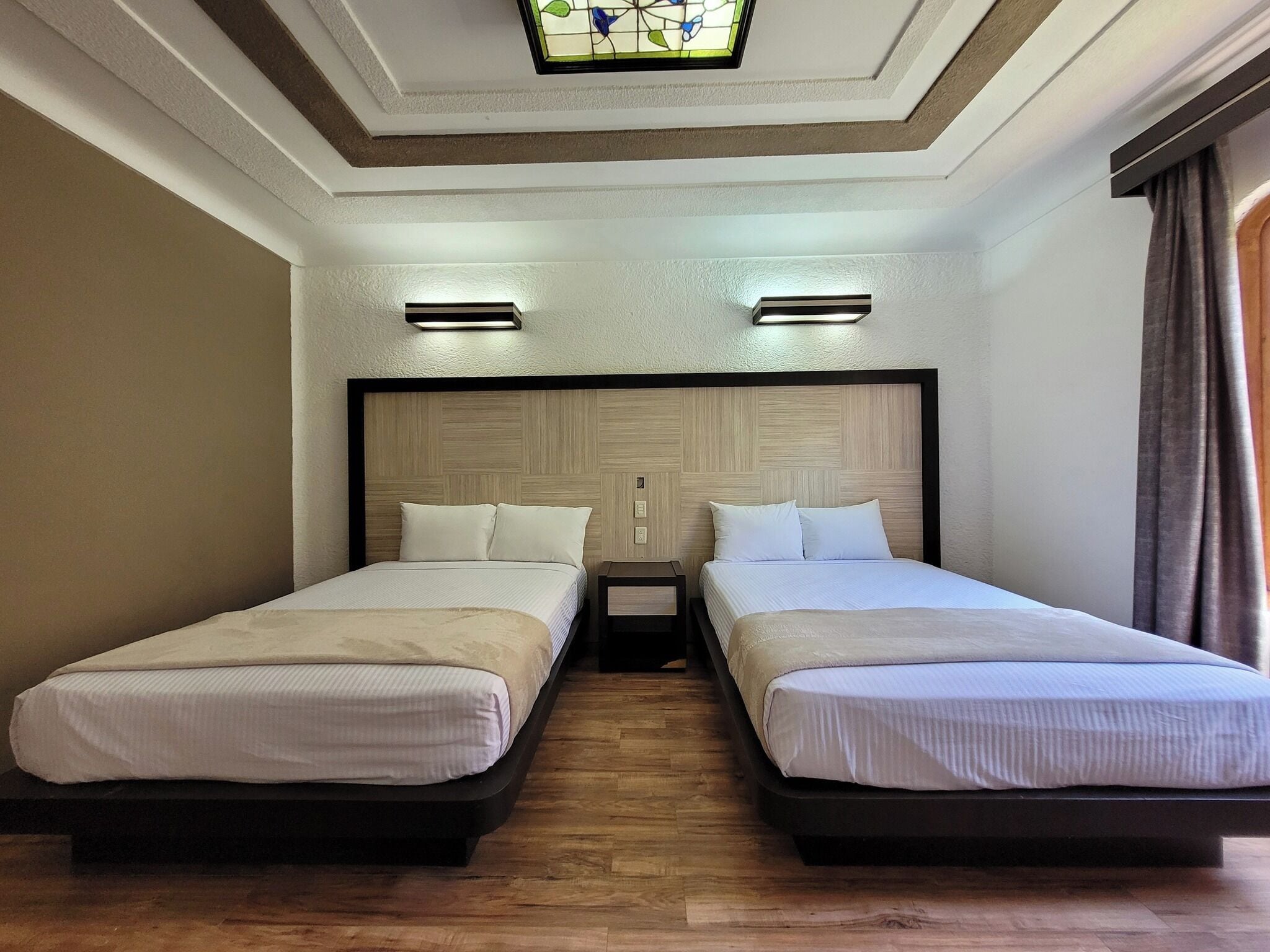 Habitación de hotel con dos camas individuales, cabecero de madera, una mesita y una lámpara en el techo.