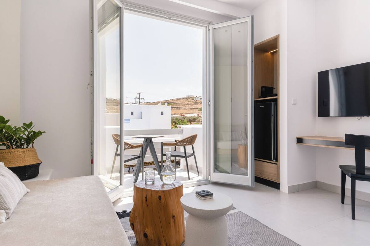 A modern bedroom with a bed, a small round table, a TV, and a sliding glass door leading to a balcony with a table and chairs.