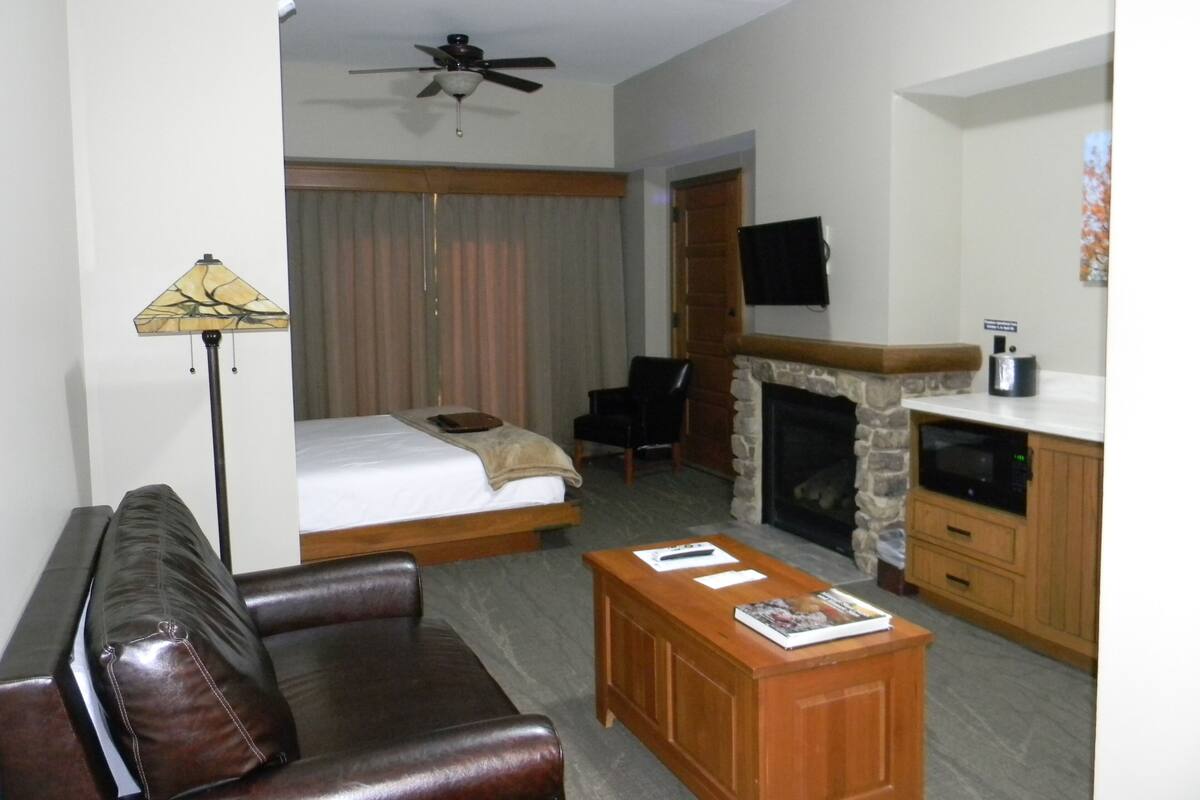 A hotel room with a bed, a wooden coffee table, a leather sofa, a fireplace, a television, and a ceiling fan.