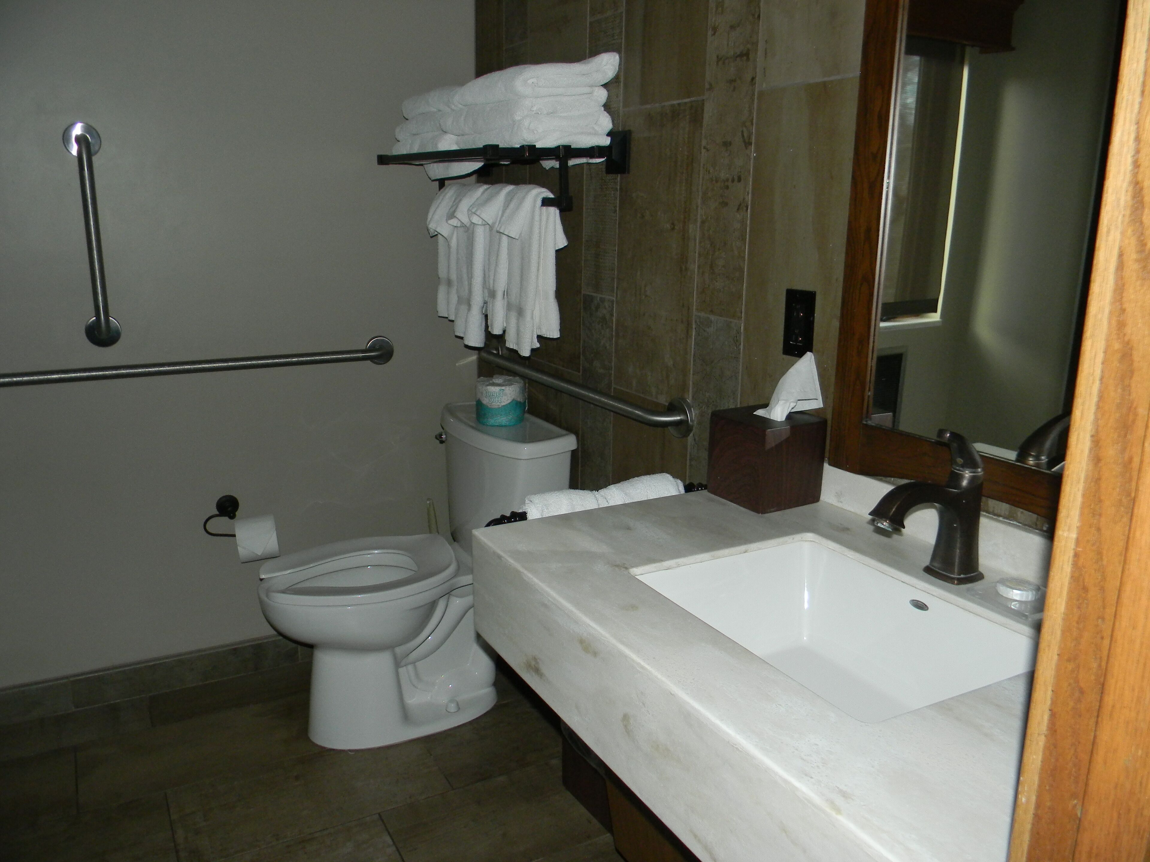 A bathroom with a white toilet, a sink with a mirror, a towel rack with folded towels, and a handrail.