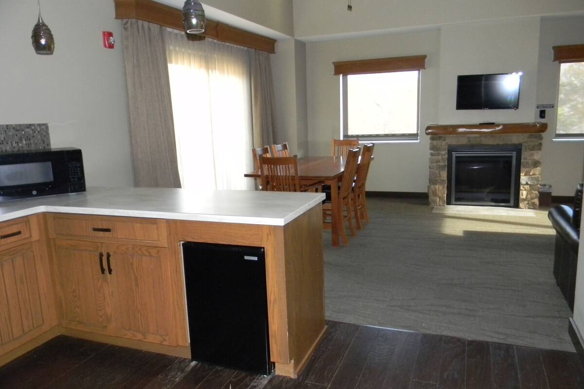 A kitchen with wooden cabinets, a built-in dishwasher, a dining area with a table and chairs, a fireplace, and a television mounted on the wall.