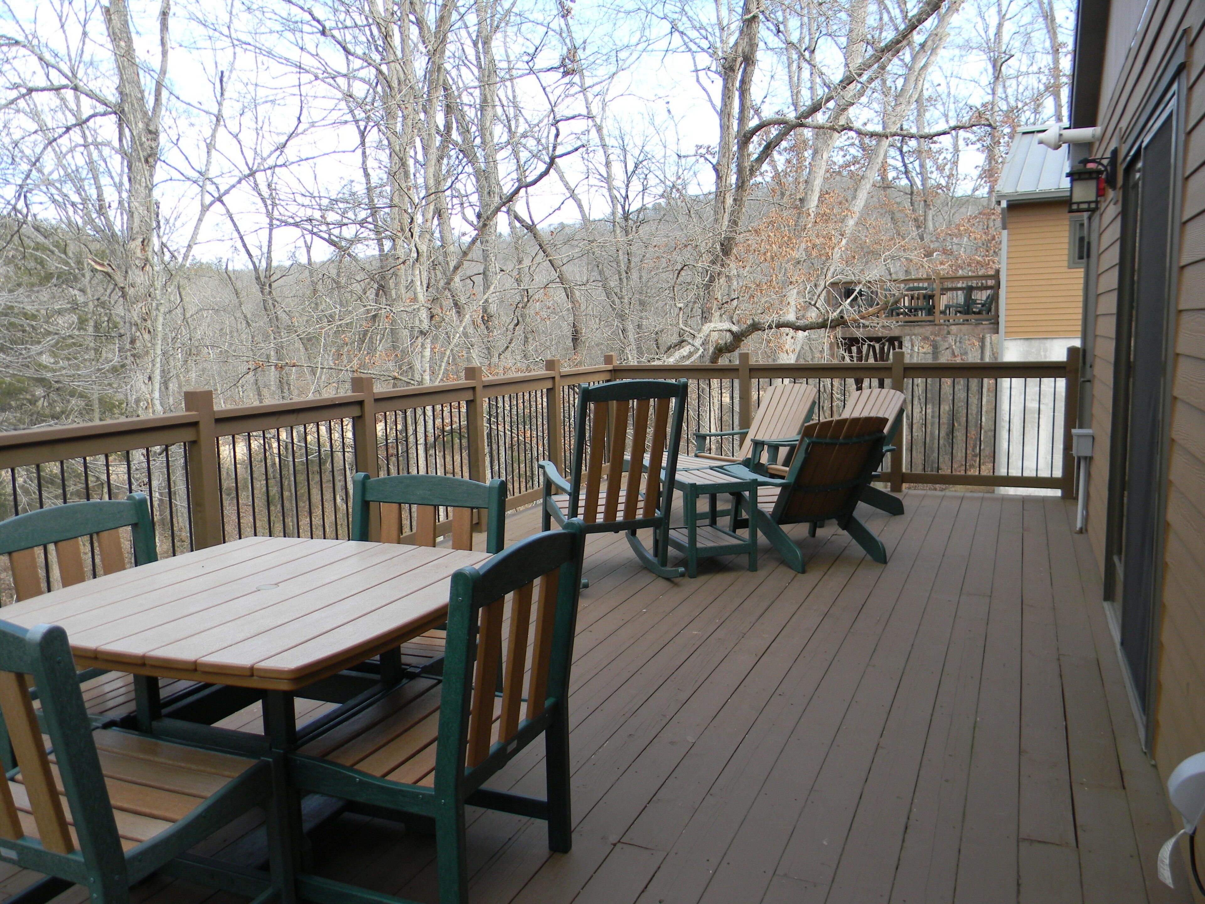 Une terrasse en bois avec une table et des chaises, une rambarde et une vue sur des arbres et un bâtiment en arrière-plan.