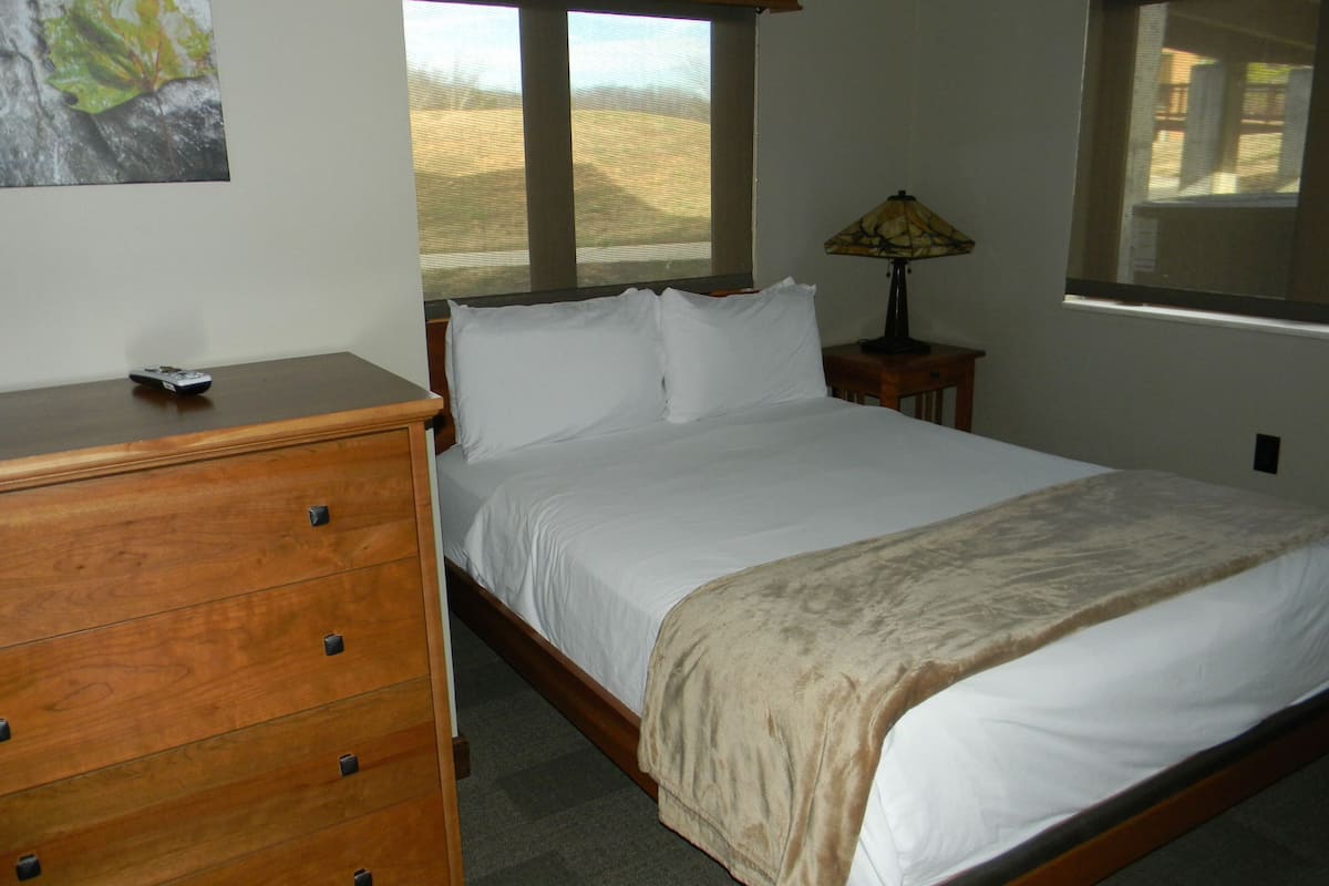 A bedroom with a bed, wooden dresser, lamp, and large windows.