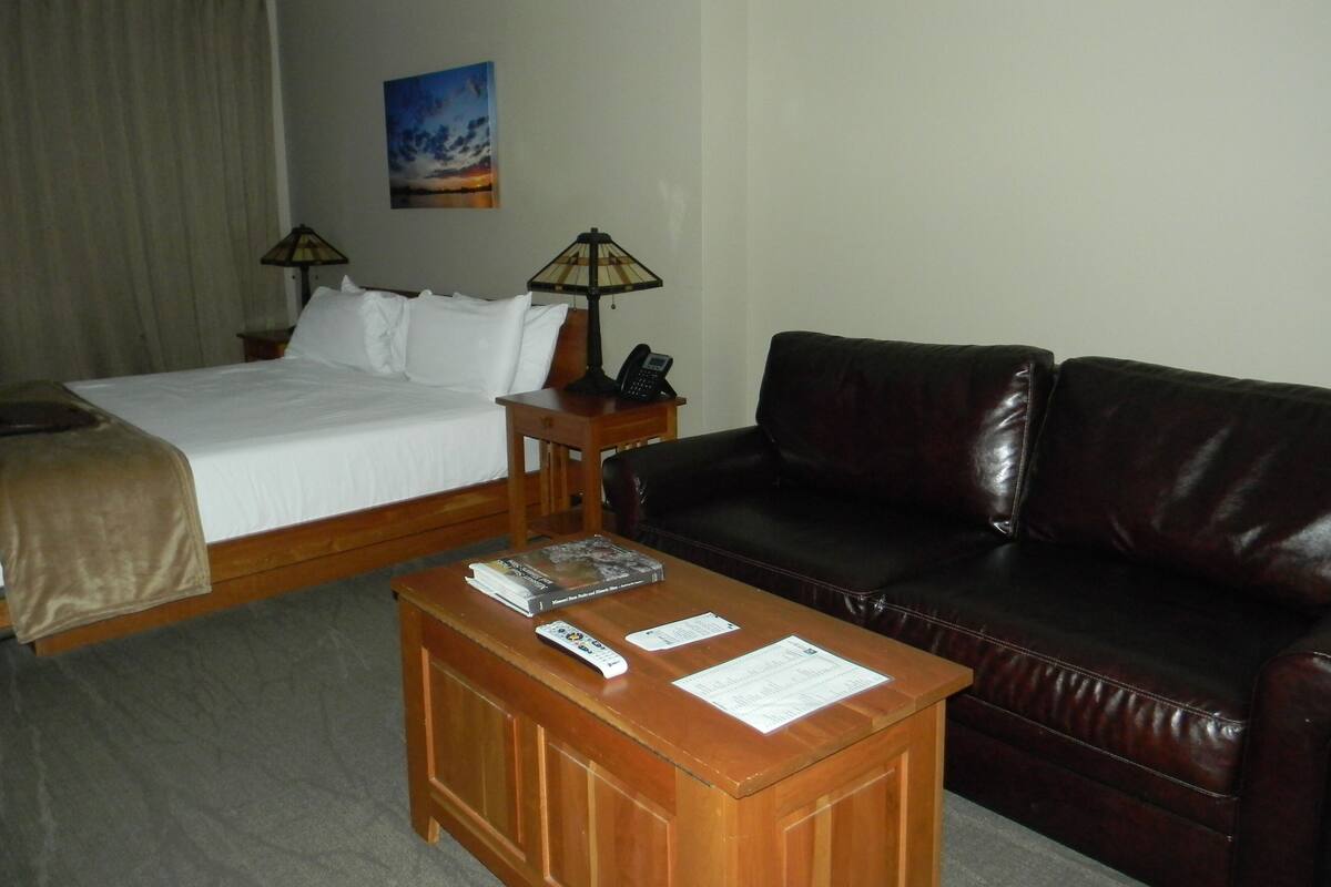A hotel room with a bed, a wooden nightstand with a lamp, a leather sofa, and a coffee table with remote controls and a menu.