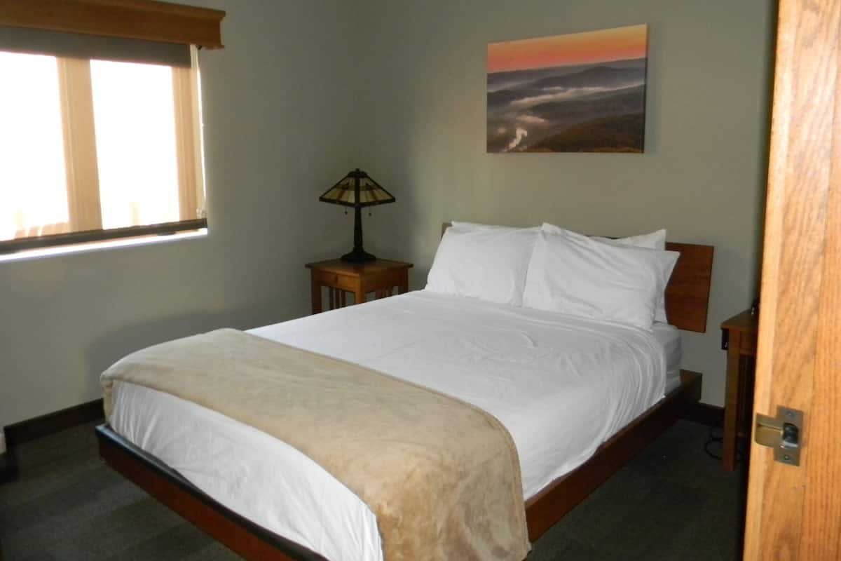 A bedroom with a bed, a nightstand, a lamp, a window, and a painting on the wall.