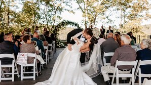 Una ceremonia de boda con invitados sentados en hileras de sillas blancas, una novia con un vestido blanco y un novio con traje negro.