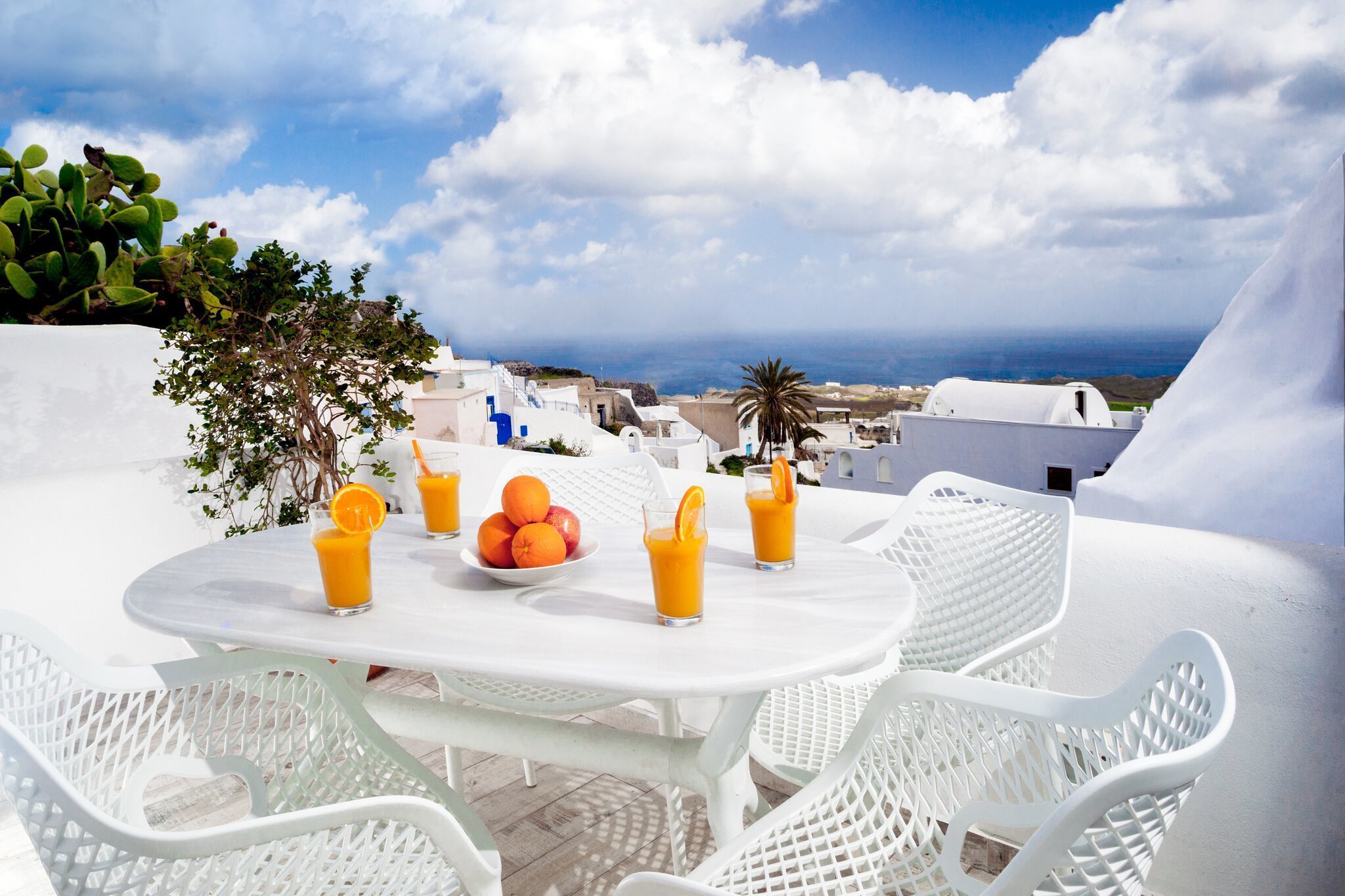 A white table with chairs, a bowl of oranges, and glasses of orange juice, overlooking a coastal town with white buildings and a view of the sea.