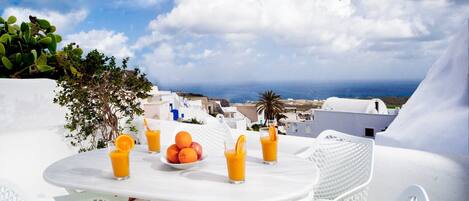 A white table with chairs, a bowl of oranges, and glasses of orange juice, overlooking a coastal town with white buildings and a view of the sea.