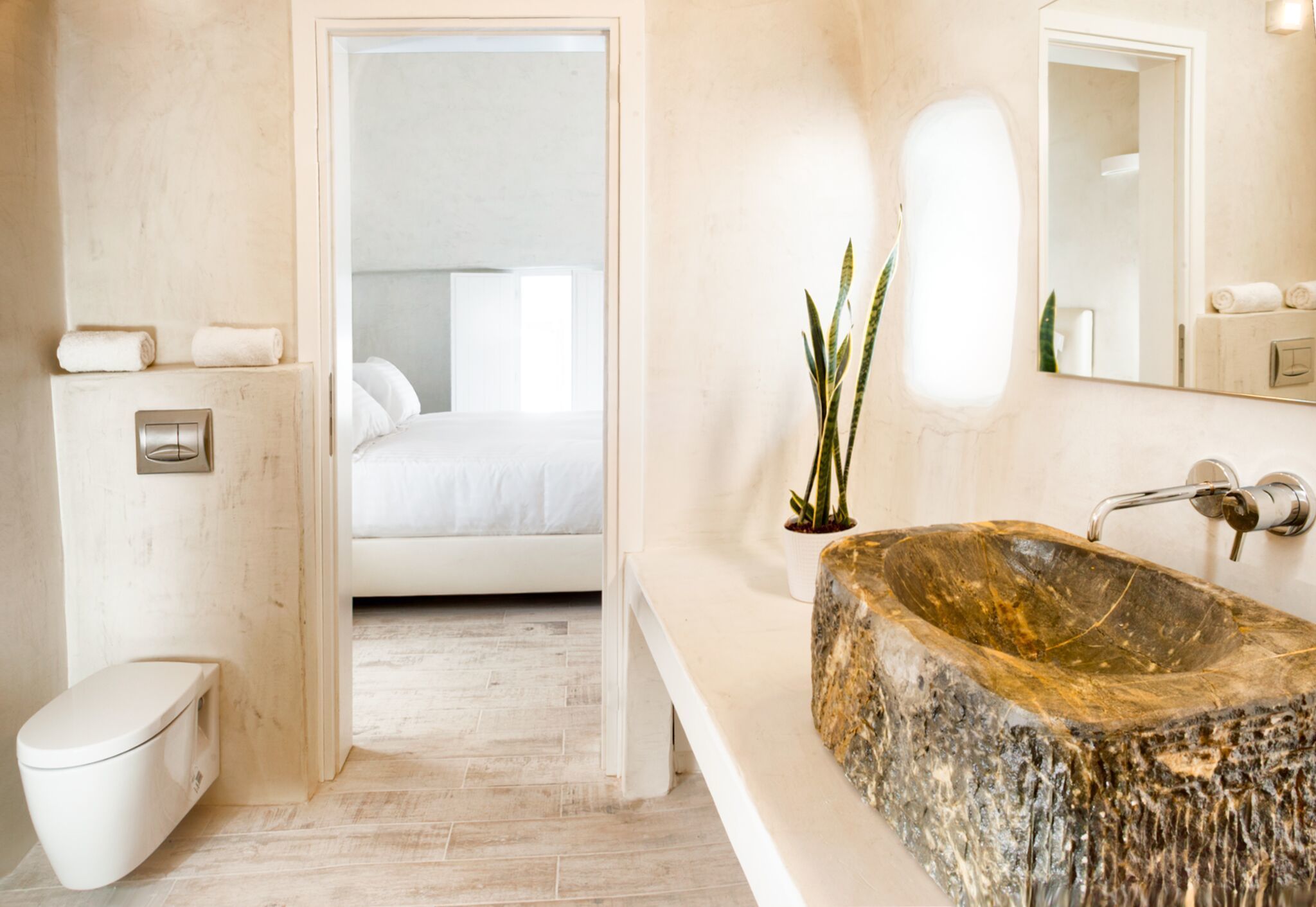 A modern bathroom with a large stone sink, a toilet, and a bed visible through an open door.