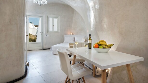 A modern, minimalist room with a white table set for two, a bowl of fruit, and a bottle of wine. - Spilies by Thireas (Santorini)