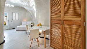 A modern bedroom with a white bed, a dining table with a bowl of fruit, and a wooden wardrobe. - Spilies by Thireas (Santorini)
