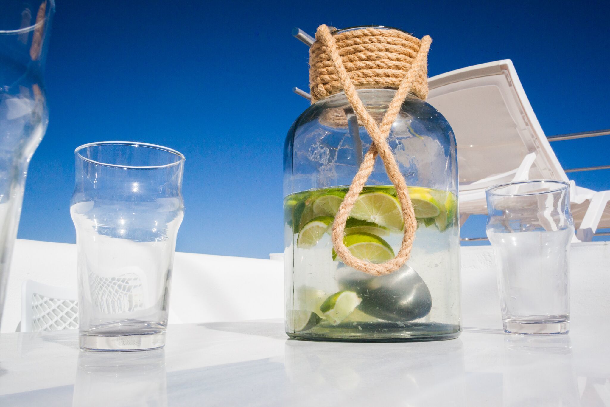 A glass jar with a rope handle filled with lime slices and ice, placed on a table with two empty glasses beside it.