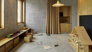 Een moderne, goed verlichte kamer met een lang houten bureau, een klein keukenhoekje en een speelruimte met speelgoed en een houten poppenhuis.