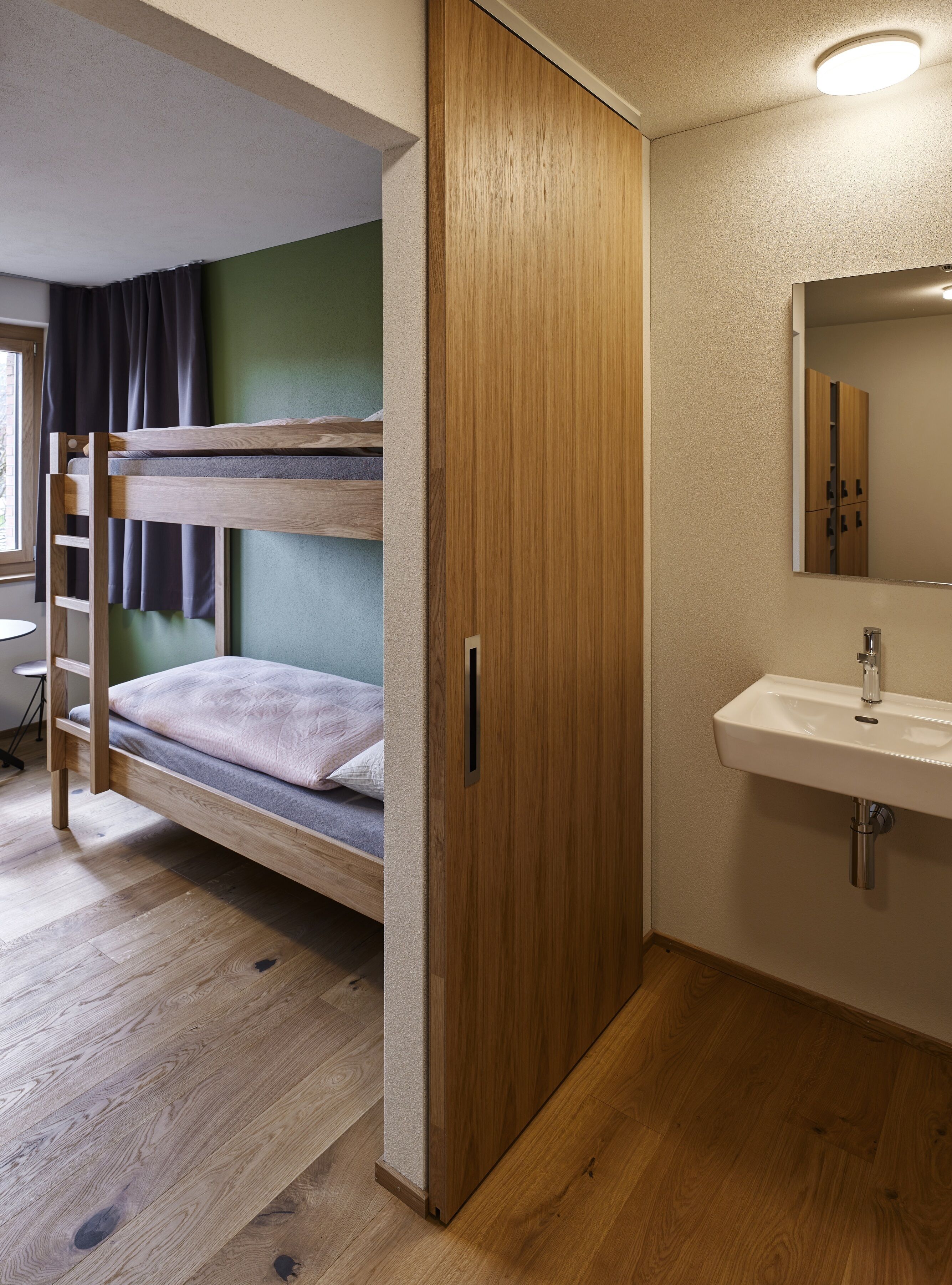 A bunk bed with a wooden ladder, a wooden wardrobe, and a bathroom with a sink and mirror.