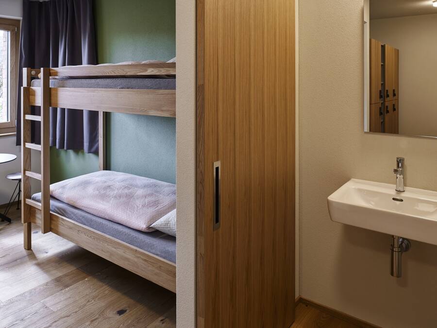 Una cama individual superpuesta con una escalera de madera, un armario de madera y un baño con lavamanos y espejo.
